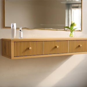 May include: A light-colored wooden floating vanity with three drawers and round knobs. The vanity has a vertical grooved design on the drawer fronts. A mirror hangs above the vanity, and two white bottles and a vase with flowers sit on top.
