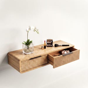 May include: A light brown wooden floating shelf with two drawers featuring vertical slats and thin black metal handles. On top sits a clear glass vase with a white orchid, a small lipstick, a compact eyeshadow palette, and a makeup brush.