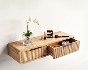 May include: A wall-mounted wooden floating shelf featuring two drawers with vertical slat detailing and thin black metal handles. A square glass vase with a white orchid, a lipstick, an eyeshadow palette, and a makeup brush sit on top.