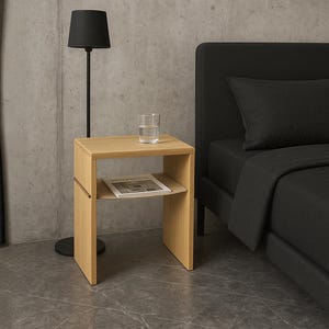 Pode incluir: Uma mesa de cabeceira em carvalho claro com uma prateleira inferior com uma revista. Um copo de água está em cima. Uma lâmpada preta e uma estrutura de cama preta estão no fundo. A mesa tem cerca de 50 cm de altura.