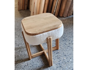 May include: A small, modern stool with a square, light-colored wooden top and a white, textured fabric skirt. The legs are made of light-colored wood and are connected by a cross-shaped support. The stool is designed for seating.