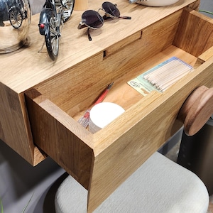 Puede incluir: Una estantería de pared de madera con un cajón abierto, que revela un pequeño recipiente de bastoncillos de algodón y un lápiz. La estantería tiene un pomo de madera y está decorada con una pequeña bicicleta, una mano de madera, gafas de sol, una botella de crema de argán y una maceta de cerámica blanca con un agujero en el lateral.