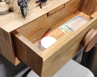 Puede incluir: Un tocador de pared de roble claro con un cajón extraíble que contiene un pequeño tarro blanco y un paquete de palitos de madera. La superficie superior sostiene un modelo de bicicleta en miniatura, una mano de madera, loción y gafas.