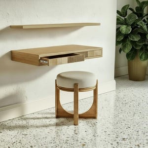 May include: A modern, light wood floating desk with a drawer and a matching shelf are mounted on a white wall. A round, upholstered stool with a wooden frame sits below. A large potted plant is in the background.