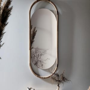 Op de afbeelding: Een ovale spiegel met een licht houten frame. Het spiegeloppervlak is zacht wit, met een zwart-wit boomafbeelding. Hij hangt aan een lichtgrijze muur. Decoratieve items staan op een houten tafel eronder.