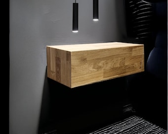 May include: A rectangular, light-toned wooden floating shelf mounted horizontally on a dark gray wall. Two slender, cylindrical black pendant lights hang vertically from the ceiling above the shelf, casting focused light downward onto the wood surface.