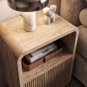 May include: A light oak bedside table with rounded corners and a fluted lower cabinet door. The top surface holds a white marble lamp and a small abstract sculpture. An open shelf contains a brown leather tray holding two stacked books.