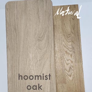 May include: Two rectangular wooden boards with visible grain patterns. The left board is labeled hoomist oak in dark text, and the right board is labeled Natural in white script. Both pieces display light brown wood tones with distinct textures.