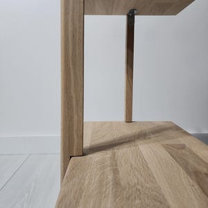 May include: Close-up of a light oak wood shelf unit. The shelves are supported by vertical wooden posts and metal brackets. The wood grain is visible on the shelves and posts. The unit is against a white wall.