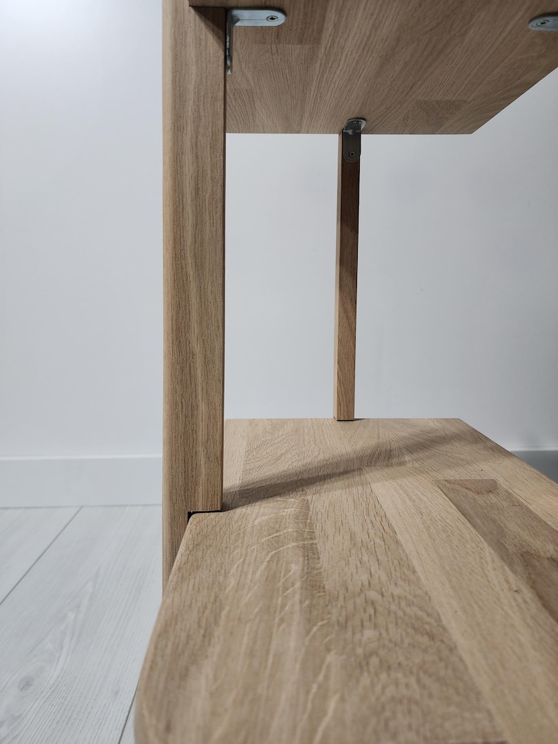 May include: Close-up of a light oak wood shelf unit. The shelves are supported by vertical wooden posts and metal brackets. The wood grain is visible on the shelves and posts. The unit is against a white wall.