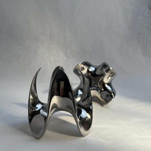 May include: Two polished silver metal objects. The first is a stylized, curved shape with the word "Sveelife" printed on it. The second is a four-lobed, abstract design. Both objects have a reflective, metallic finish and are set against a white background.