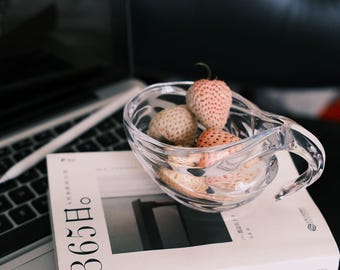 May include: A clear glass bowl with a handle, filled with several strawberries, sits atop a white book with the text "365日." The book rests on a laptop keyboard. The strawberries are light pink and beige.