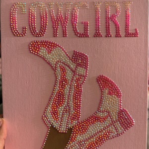 May include: Pink canvas with the word "COWGIRL" in pink rhinestones.  Two brown legs wearing pink and white rhinestone cowboy boots.
