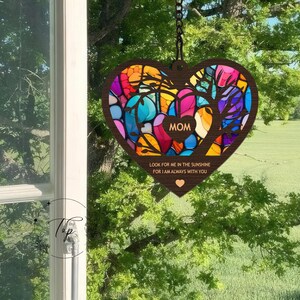 May include: A wooden heart-shaped wind chime with a stained glass design in shades of blue, green, red, and yellow. The heart has the word "MOM" in the center and the text "Look for me in the sunshine for I am always with you".