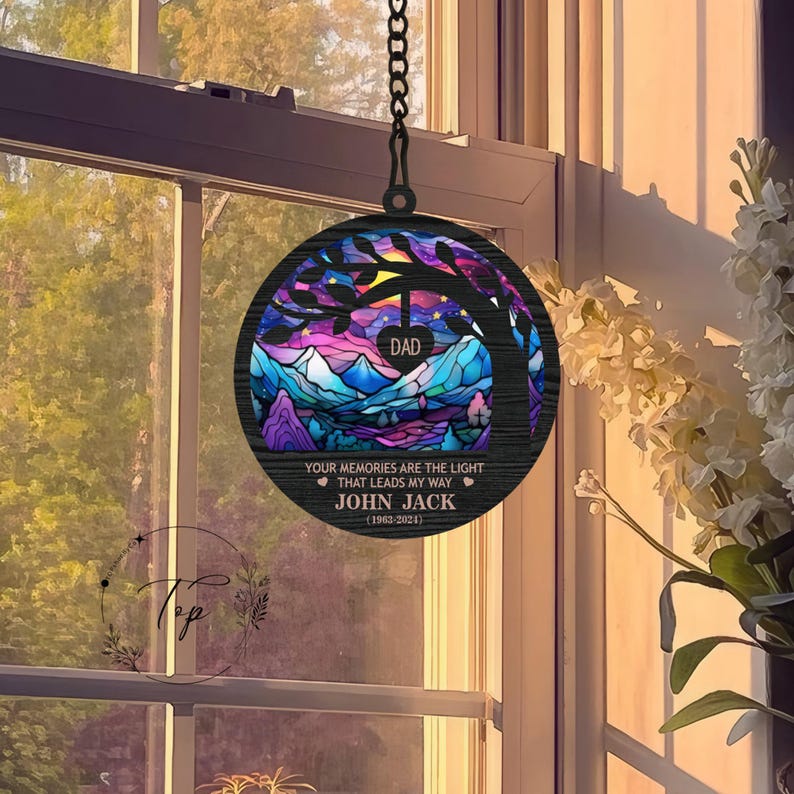 May include: A wooden stained glass style memorial wind chime with a tree and mountain landscape. The chime has the text "DAD", "YOUR MEMORIES ARE THE LIGHT THAT LEADS MY WAY", "JOHN JACK", and "(1963-2024)".