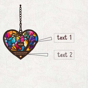 May include: A colorful stained glass heart-shaped ornament with a chain for hanging. The ornament features a tree design and the word "MOM" in the center. The text "LOOK FOR ME IN THE SUNSHINE FOR I AM ALWAYS WITH YOU" is written below the tree.