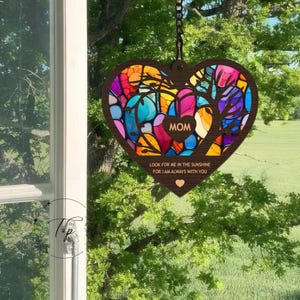 May include: A wooden heart-shaped wind chime with a stained glass design in shades of blue, green, red, and yellow. The heart has the word "MOM" in the center and the text "Look for me in the sunshine for I am always with you".