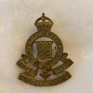 Puede incluir: Una insignia de metal dorado con las palabras "Royal Canadian Ordnance Corps" y una corona en la parte superior.