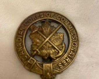 May include: A gold-coloured metal pin with the words "Canadian Corps of Commissioners" around the edge. The centre of the pin features a maple leaf with crossed swords and an anchor.