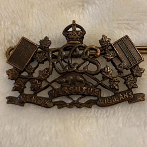 Op de afbeelding: Een vintage metalen pin met een kroon, esdoornbladeren en de letters RCS. De pin heeft ook de woorden "Velox", "Versutus" en "Vigilans" op een banner.