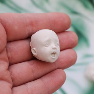 May include: A small, unpainted doll head with an open mouth and surprised expression. The doll head is held in a person's hand, showcasing the fine details of the sculpted features.