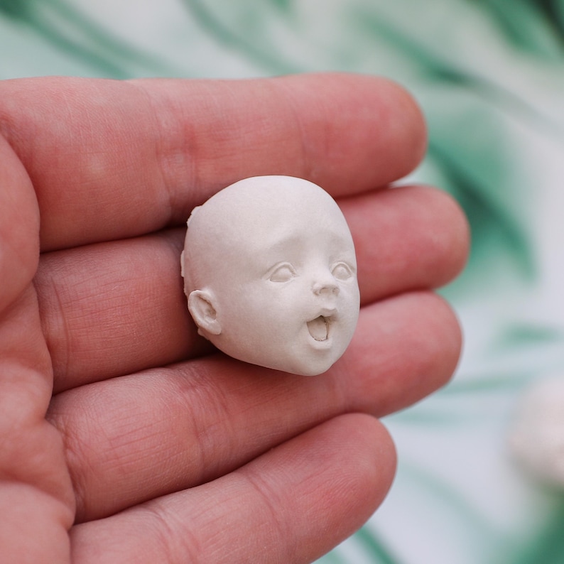 May include: A small, unpainted doll head with an open mouth and surprised expression. The doll head is held in a person's hand, showcasing the fine details of the sculpted features.
