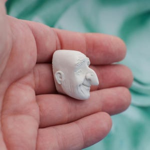 May include: A small, off-white sculpture of a smiling man's head. The sculpture has detailed features, including a prominent nose, a receding hairline, and a visible ear. The piece is held in a person's hand, with a blurred turquoise background.