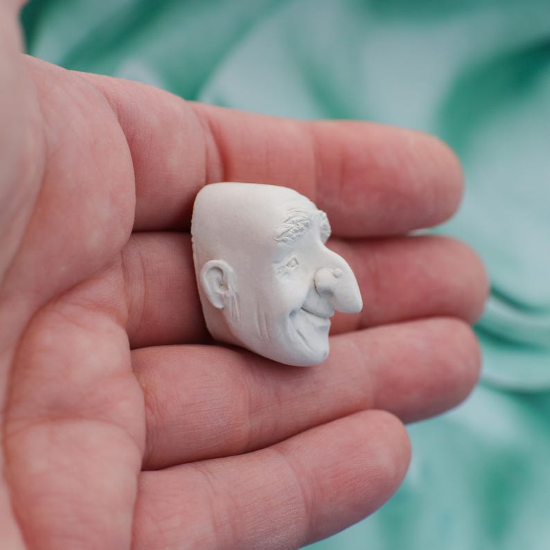 May include: A small, off-white sculpture of a smiling man's head. The sculpture has detailed features, including a prominent nose, a receding hairline, and a visible ear. The piece is held in a person's hand, with a blurred turquoise background.