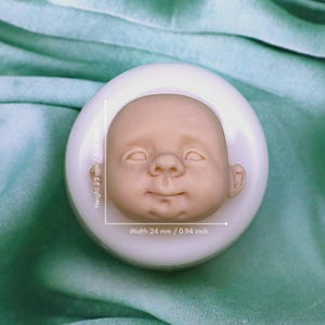 Könnte beinhalten: Eine Silikonform zum Herstellen eines Baby-Puppenkopfes. Die Form ist weiß und hat ein Babygesicht, das hineingeschnitzt ist. Die Form ist 2,5 Zoll hoch und 0,94 Zoll breit.