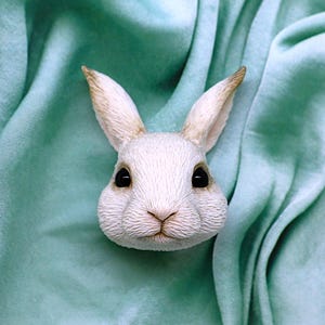 Può includere: Una scultura a forma di testa di coniglio bianca con una texture di pelliccia dettagliata e grandi occhi neri. Il coniglio ha lunghe orecchie con punte marroni e un naso piccolo e dettagliato. La scultura è ambientata su uno sfondo di tessuto verde chiaro e morbido.