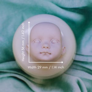 May include: A pale, unpainted doll head, 1.42 inches tall and 1.14 inches wide, rests on a circular base. The doll head has closed eyes, a small nose, and a slightly parted mouth. The background is a soft, green fabric.