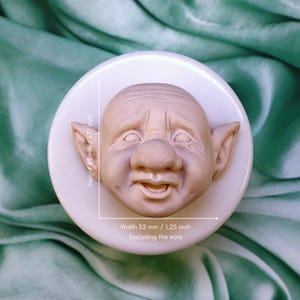 Peut inclure: Un visage sculpté beige clair avec un grand nez et de grandes oreilles, monté sur une base circulaire blanche. Le visage mesure 45 mm de haut et 32 mm de large, sans les oreilles. Le visage a une expression souriante.