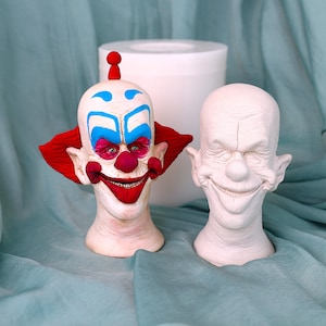 Könnte beinhalten: Zwei Clownkopf-Skulpturen. Eine ist mit roten, blauen und rosa Details bemalt, einschließlich eines roten Pompons oben. Die andere ist eine leere, unbemalte Skulptur mit einem breiten Grinsen. Beide stehen auf einer hellblauen Oberfläche.