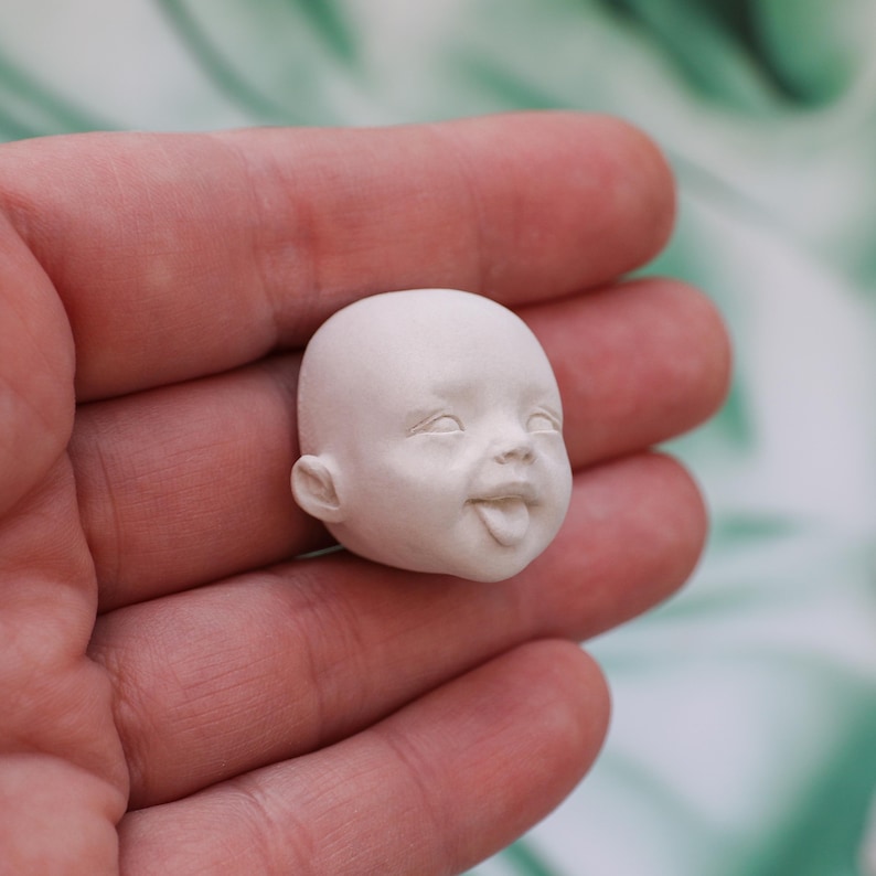 May include: A small, unpainted, white doll head with a smiling expression and tongue sticking out. The doll head has detailed facial features, including eyes, ears, and a smooth surface.