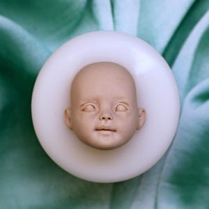 May include: A silicone mold for making a doll head. The mold is white and has a detailed impression of a baby's face.
