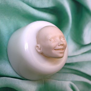 Peut inclure: Une sculpture de tête humaine souriante et pâle émergeant d'une forme arrondie blanche. La tête présente des traits faciaux détaillés et se détache sur un fond doux de couleur sarcelle. La sculpture semble être faite d'un matériau lisse et mat.