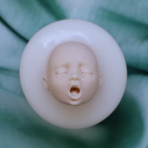 May include: A pale, sculpted baby head with closed eyes and an open mouth, set within a white, circular mold. The head is centered, with fine details visible. The background is a blurred teal color.