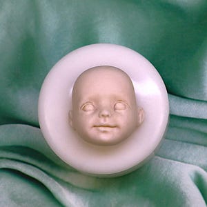 Puede incluir: Una cabeza de muñeca pálida, de piel lisa, con los ojos cerrados y una ligera sonrisa, colocada en un molde circular blanco. La cabeza de la muñeca está centrada sobre un fondo de tela verde, creando una estética suave y serena.