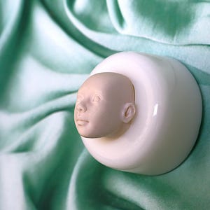 Pu&ograve; includere: Uno stampo in ceramica bianca con un viso di bambino scolpito al suo interno. Lo stampo &egrave; su uno sfondo di tessuto verde chiaro.