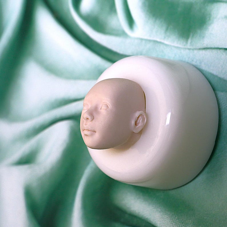 Pu&ograve; includere: Uno stampo in ceramica bianca con un viso di bambino scolpito al suo interno. Lo stampo &egrave; su uno sfondo di tessuto verde chiaro.