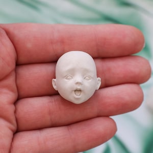 May include: A small, off-white doll head with a surprised expression. The head is bald, with detailed facial features including open eyes and a slightly open mouth. The doll head is held in a hand.