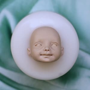 Puede incluir: Un molde de silicona para crear una cabeza de muñeca bebé. El molde es blanco y tiene una impresión detallada de la cara de un bebé con los ojos cerrados y una leve sonrisa.