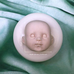 Può includere: Uno stampo circolare in silicone bianco con un volto di neonato dettagliato. Lo stampo ha un'altezza di 25 mm e una larghezza di 20 mm, come indicato dalle linee di misurazione bianche stampate direttamente sopra l'oggetto.