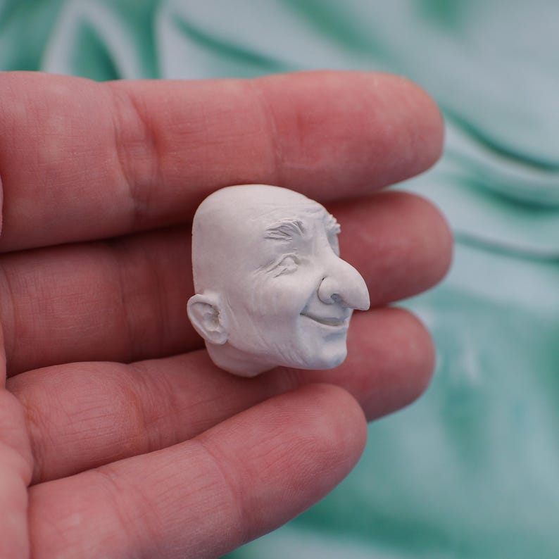 May include: A small, white sculpture of a smiling man's head. The sculpture has a prominent nose, detailed wrinkles, and a bald head. The figure is held in a person's hand, with a blurred background.
