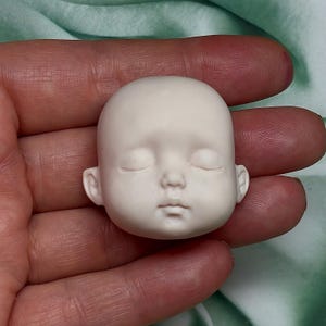 May include: A small, unpainted doll head with closed eyes, a small nose, and a defined mouth. The doll head is a light beige color and is held in a hand against a soft green background. The head is smooth and appears to be made of a hard material.