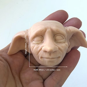 May include: A detailed sculpture of a character's face, with closed eyes and large ears. The face is a light beige color, with visible wrinkles and fine details. The height is 2 inches, and the width is 1.53 inches plus ears.