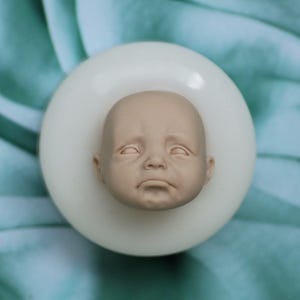 Puede incluir: Un molde de silicona blanco para hacer una cabeza de muñeca de bebé. El molde tiene la forma de la cara de un bebé con los ojos cerrados y una expresión ligeramente triste.