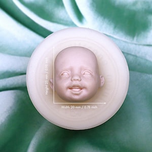 May include: A pale baby doll head, 2.5 cm tall and 2 cm wide, set in a round, off-white mould. The doll head has closed eyes and a slight smile. The background is a soft, green fabric.