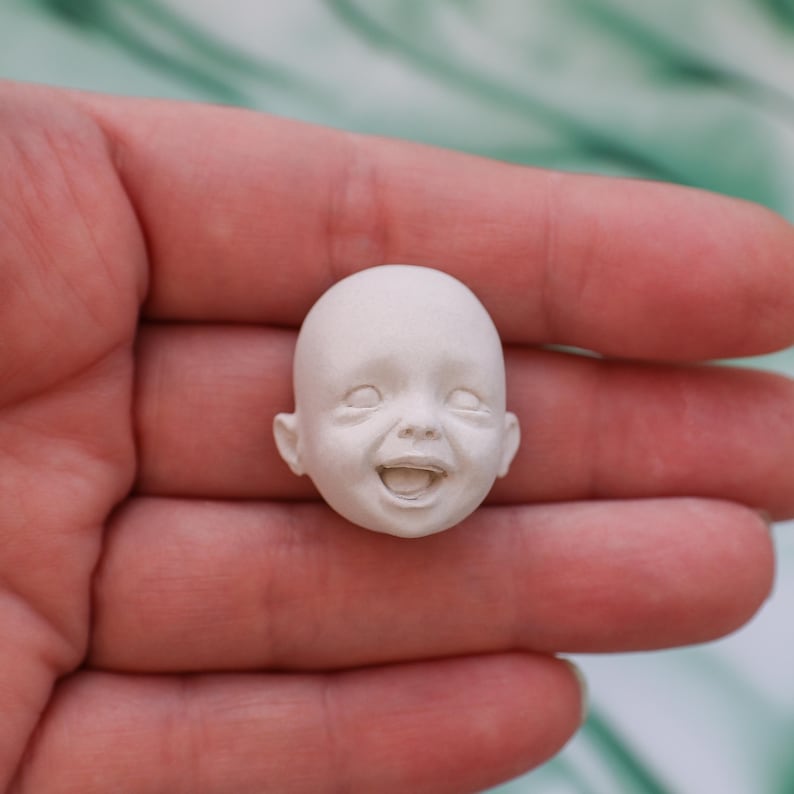 May include: A small, off-white, smiling baby doll head. The doll head has closed eyes and an open mouth, revealing a detailed sculpt of the teeth and tongue. The head is held in a person's hand, with a blurred green background.