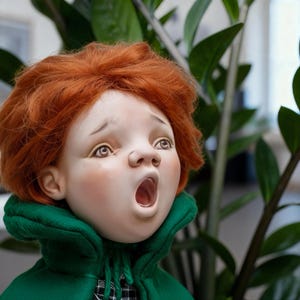 Puede incluir: Muñeca de porcelana con pelo rojo y abrigo verde. La muñeca tiene una expresión de sorpresa con la boca abierta. La cara de la muñeca tiene detalles, incluyendo ojos pintados y mejillas rosadas. Una pieza de colección única.
